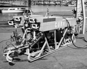 A black and white photograph of a boxy remotely operated vehicle with a grasping claw at the front