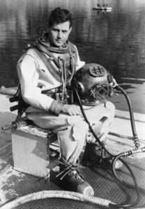 Navy diver sits on box while wearing a historic diving suit and holding a diving helmet in his lap