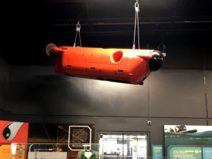 A large orange-red remotely operated vehicle is suspended from the ceiling of an exhibit gallery