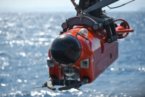 Front view of mine neutralization vehicle suspended over water. Vehicle is bright orange/red-colored with a hemispherical black dome on the nose.