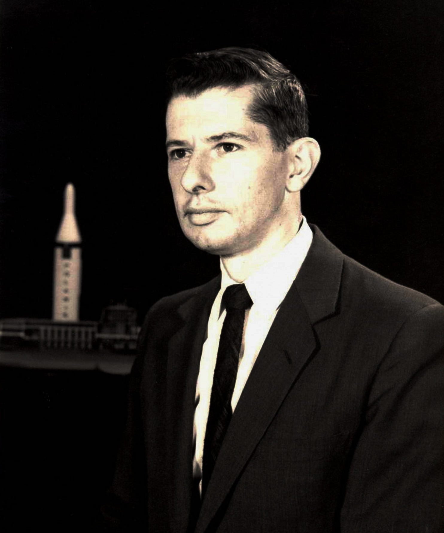 A young man looks away from the camera with a neutral expression on his face. Small models of a Polaris missile and a submarine sit in the background.