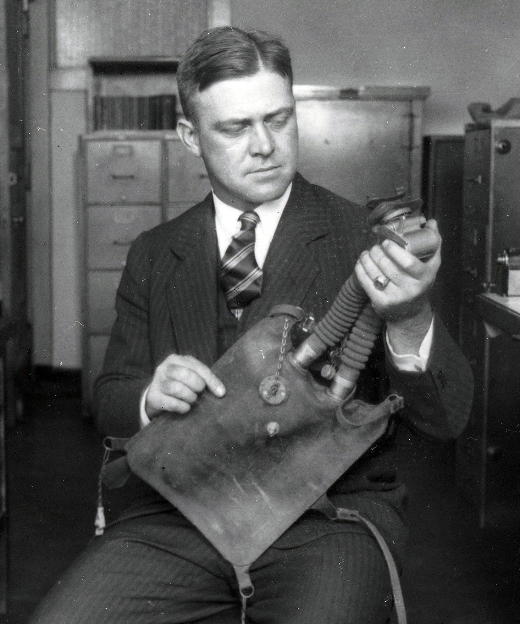 Man sitting in a chair holding a submarine escape device which looks like a boxy canvas bag with breathing hose