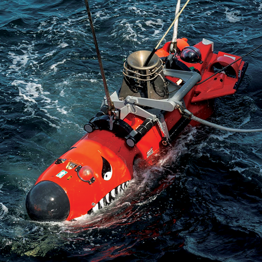 A red-orange remotely operated vehicle is lowered into the water. Decals with eyes and pointy teeth are attached to the front of the vehicle.