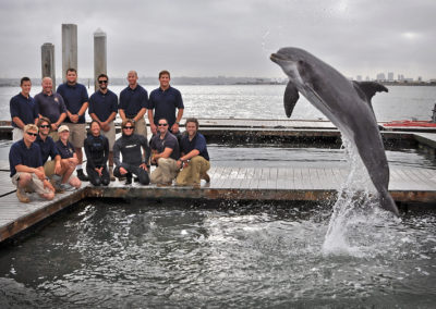 A dolphin jumps out of the water as a group of people pose in the background