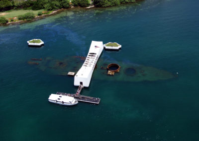 The rusted wreckage of battleship Arizona lies under blue water. A white memorial structure sits perpendicularly above the wreckage.