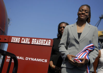 A young woman holds a ribbon-wrapped christening bottle. She stands next to a red rudder with text reading "USNS Carl Brashear General Dynamics."
