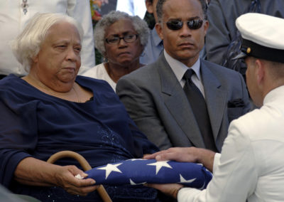 A Sailor hands a folded flag to an older woman.