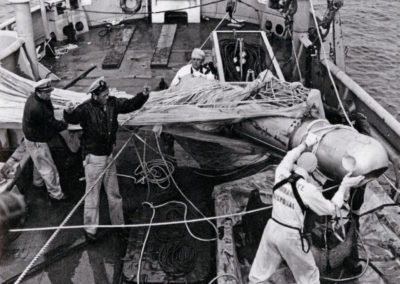 Sailors bring a damaged hydrogen bomb onto the deck of a ship. A parachute is tangled around the bomb.