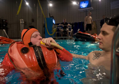 A submariner in a bright red-orange escape suit blows into a tube attached to the suit's arm.