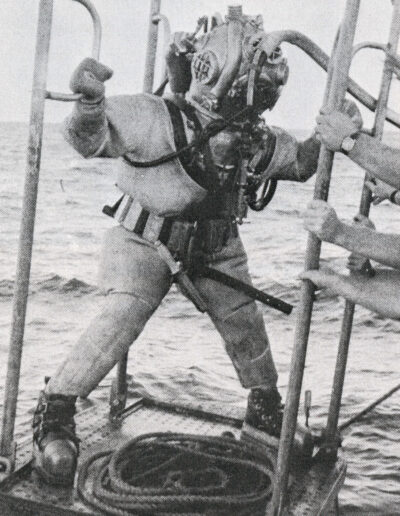 A diver wearing ta Mark 5 helium diving rig stands on a diving stage as several arms push the stage towards the water.