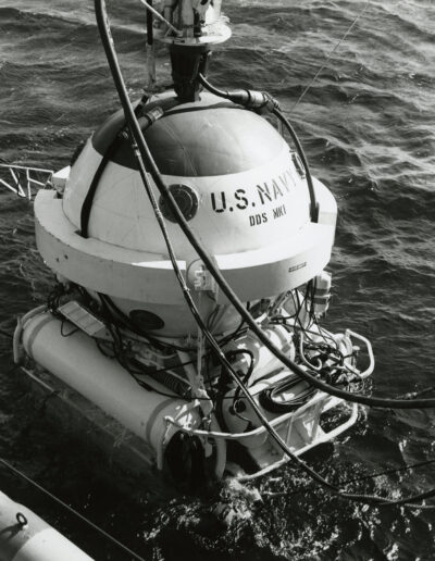 A white sphere with cables and cylinders is lowered into the water. Stenciled on the sphere is "U.S. Navy DDS Mark 1."