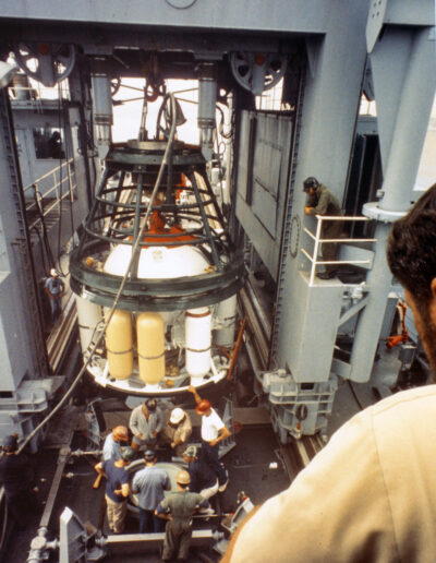 A white and yellow transfer capsule is suspended inside a ship well. People in hard hats stand underneath it.