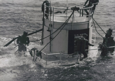 Divers climb around the Sealab II habitat as it's lowered below the water's surface.