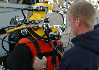 A diver wears a red and black hot water suit and a yellow Mark 21 helmet with several cables.
