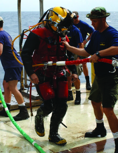 A diver wears a red and black hot water suit and a yellow Mark 21 diving helmet.