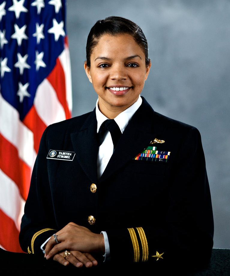 A young female African American Navy officer smiles at the camera. She wears a Navy dress uniform and poses before an American flag.