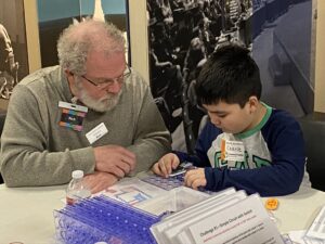 A Discover E Day and museum volunteer sits at a table helping a young museum visitor build an electric snap-together circuit at E Day 2020.