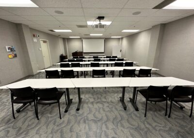 View of room from back at entry door, looking towards front, with rows of white tables and black chairs. Projector screen visible on front wall.