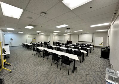 Back of room looking towards front, rows of white tables and black chairs facing front. Narrow white tables at room edges. Whiteboards around edges of the room. Lights on. Two projector screens at front of room hanging from ceiling. Lectern in front center of room.